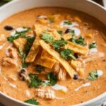 Creamy chicken tortilla soup in white bowl topped with cheese, cilantro, and tortilla strips