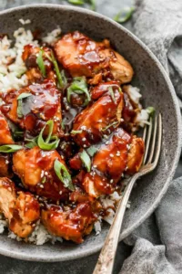 Crock pot bourbon chicken served over brown rice and topped with chopped green onions in a white bowl