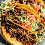 Crispy ground beef tacos in homemade fried corn tortilla shells topped with shredded lettuce, tomatoes, and cheddar cheese