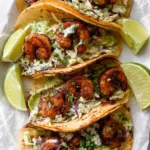 Four grilled shrimp tacos with avocado lime slaw and sliced avocado on a parchment-lined cutting board