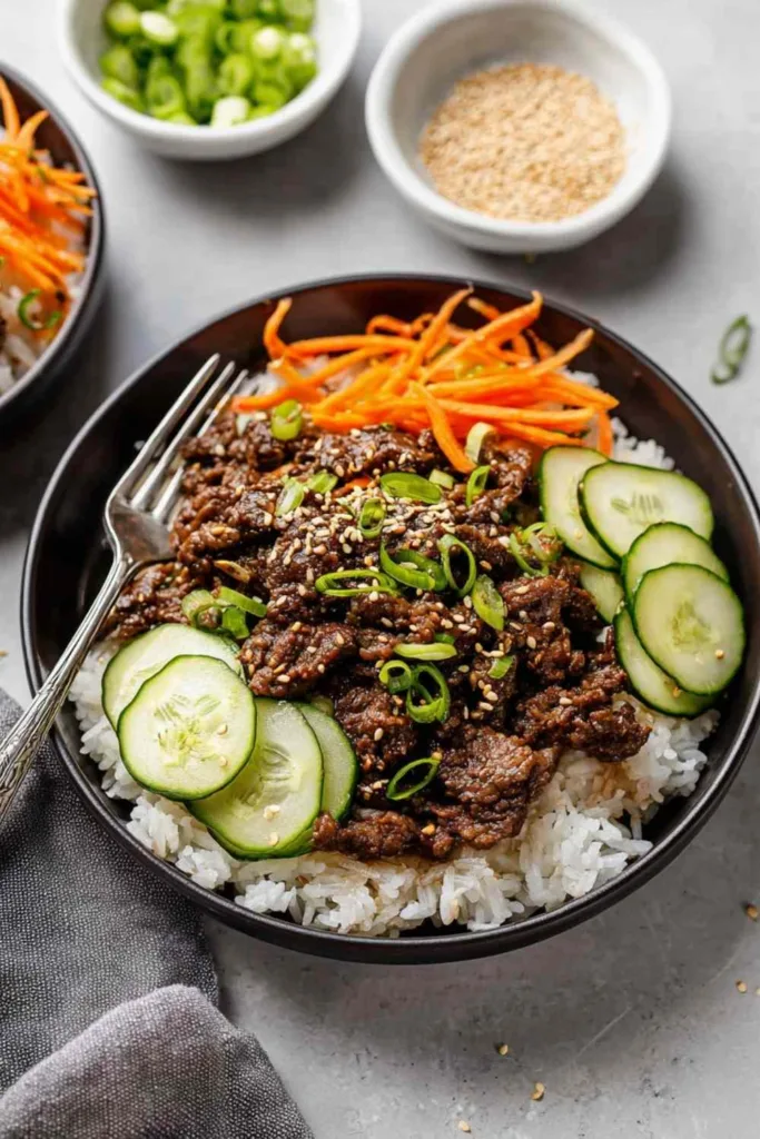 Korean ground beef rice bowl topped with cucumber salad and sliced green onions in a white bowl