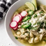 Overhead view of slow cooker pozole verde in a white bowl topped with avocado cilantro and lime