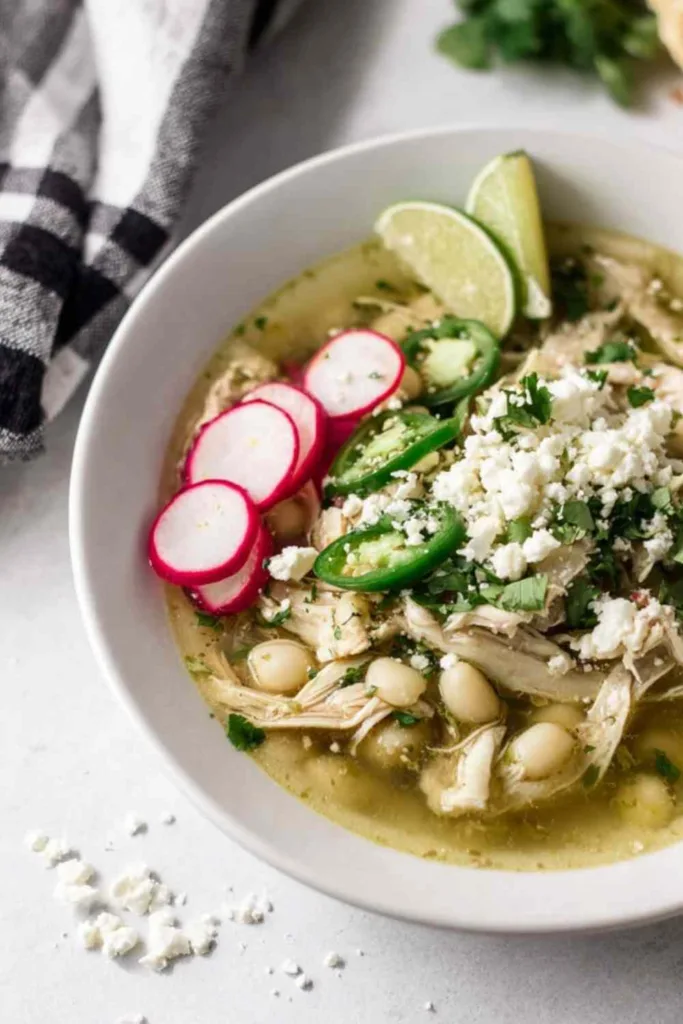 Overhead view of slow cooker pozole verde in a white bowl topped with avocado cilantro and lime