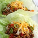Ground turkey taco lettuce wraps served on a white plate topped with pico de gallo and shredded cheddar