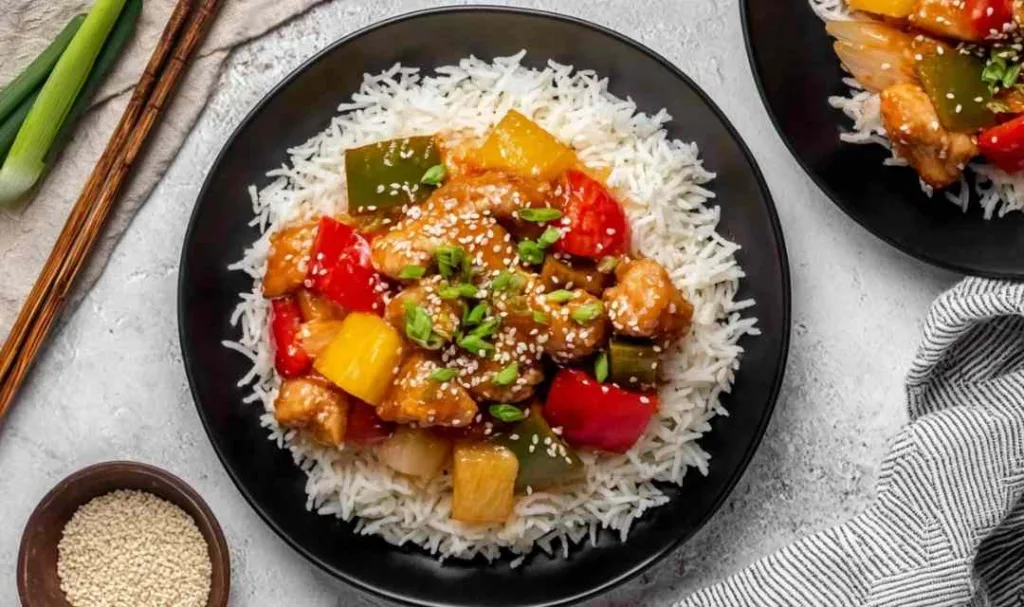 Slow cooker sweet and sour chicken served over white rice with bell peppers and pineapple garnished with green onions and sesame seeds