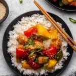 Slow cooker sweet and sour chicken served over white rice with bell peppers and pineapple garnished with green onions and sesame seeds